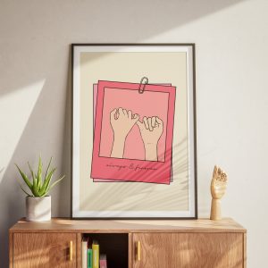 A framed poster with a minimalistic design featuring two hands making a pinky promise, set against a beige background with pink borders. The text "always & forever" is written at the bottom. The poster is placed on a wooden cabinet with a potted plant and a wooden hand sculpture, bathed in natural sunlight.