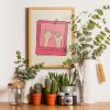 A framed poster with a minimalistic design featuring two hands making a pinky promise, set against a beige background with pink borders. The text "always & forever" is written at the bottom. The poster is surrounded by green plants and placed on a wooden shelf.