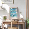 A framed poster with bold blue text reading "DON'T LOOK BACK" in all caps, with the words "you're not going that way" in smaller blue text below. The poster is displayed on a wooden desk with various items, under natural sunlight.