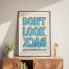 A framed poster with bold blue text reading "DON'T LOOK BACK" in all caps, with the words "you're not going that way" in smaller blue text below. The poster is placed on a wooden cabinet with a potted plant and a wooden hand sculpture, bathed in natural sunlight.