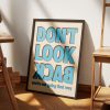 A framed poster with bold blue text reading "DON'T LOOK BACK" in all caps, with the words "you're not going that way" in smaller blue text below. The poster is leaning against a white wall on a wooden floor, illuminated by soft natural light, with parts of wooden furniture visible around.