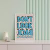 A framed poster with bold blue text reading "DON'T LOOK BACK" in all caps, with the words "you're not going that way" in smaller blue text below. The poster is placed on a mint green wall shelf next to a decorative vase and a glass.
