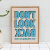 A framed poster with bold blue text reading "DON'T LOOK BACK" in all caps, with the words "you're not going that way" in smaller blue text below. The poster is leaning against a white tiled wall, placed next to a potted plant.