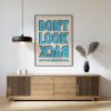 A framed poster with bold blue text reading "DON'T LOOK BACK" in all caps, with the words "you're not going that way" in smaller blue text below. The poster is hung above a wooden sideboard with decorative items and a modern pendant light.