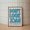 A framed poster with bold blue text reading "DON'T LOOK BACK" in all caps, with the words "you're not going that way" in smaller blue text below. The poster is leaning against a tiled wall with a light wood frame, placed on the floor.