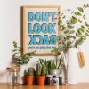 A framed poster with bold blue text reading "DON'T LOOK BACK" in all caps, with the words "you're not going that way" in smaller blue text below. The poster is surrounded by green plants and placed on a wooden shelf.