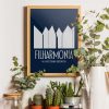 A navy blue framed poster with a minimalist design of abstract building shapes and the text "FILHARMONIA IM. MIECZYSŁAWA KARŁOWICZA" in white letters is displayed on a wooden surface against a white wall. The setting includes various potted plants, a glass jar with a wooden lid, a vintage camera, and glass bottles, creating a vibrant and eclectic display.