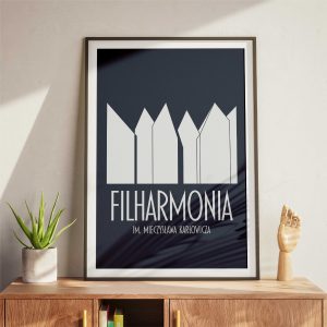 A navy blue framed poster with a minimalist design of abstract building shapes and the text "FILHARMONIA IM. MIECZYSŁAWA KARŁOWICZA" in white letters leans against a white wall above a wooden sideboard. The setup includes a small plant and a decorative wooden hand.