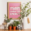 A pink framed poster with the phrase "MAIN CHARACTER ENERGY" in bold white letters is displayed on a wooden surface against a white wall. The setting includes various potted plants, a glass jar with a wooden lid, a vintage camera, and glass bottles, creating a vibrant and eclectic display.