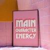 A pink framed poster with the phrase "MAIN CHARACTER ENERGY" in bold white letters is leaning against a pink tiled wall. The area around the poster features a combination of pink and yellow tiles, adding a playful and colorful atmosphere to the scene.