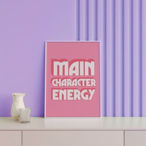 A pink framed poster with the phrase "MAIN CHARACTER ENERGY" in bold white letters is displayed against a lavender wall with vertical grooves, accompanied by a decorative white vase and a glass candle holder on a white sideboard.