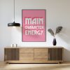 A pink framed poster with the phrase "MAIN CHARACTER ENERGY" in bold white letters is mounted on a light grey wall above a wooden sideboard. The setup includes a black pendant lamp, two white candles, and a vase with dried plants.