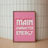A pink framed poster with the phrase "MAIN CHARACTER ENERGY" in bold white letters leans against a tiled wall. The poster is placed on a light peach-colored floor.