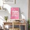 A pink framed poster with the phrase "MAIN CHARACTER ENERGY" in bold white letters is positioned on a wooden desk in a brightly lit room. The desk is decorated with a lamp, books, and a small plant, with a white wall and framed artwork in the background.