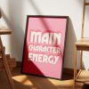 A pink framed poster with the phrase "MAIN CHARACTER ENERGY" in bold white letters leans against a white wall, resting on wooden flooring, with the shadow of a window casting across the poster.