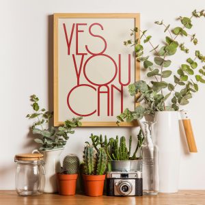 A framed beige poster with red text reading "YES YOU CAN" in a modern, minimalist font is displayed on a wooden table. The setup includes various potted plants, glass bottles, a vintage camera, and a white ceramic jug, creating a fresh and motivational arrangement with a touch of greenery.