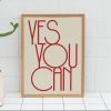 A beige framed poster with red text reading "YES YOU CAN" in a modern, minimalist font is leaning against a white tiled wall. The poster is accompanied by a small potted cactus on the floor, adding a touch of greenery to the scene.