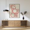 A beige framed poster with red text reading "YES YOU CAN" in a modern, minimalist font is displayed on a white wall above a wooden sideboard. The sideboard features a black hanging lamp, a black vase with dried plants, and white decorative items, creating a stylish and motivational setup.