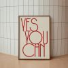 A beige framed poster with red text reading "YES YOU CAN" in a modern, minimalist font is leaning against a white tiled wall. The poster rests on a light beige floor, creating a simple and clean presentation.
