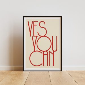 A beige framed poster with red text reading "YES YOU CAN" in a modern, minimalist font is displayed against a plain white wall. The poster rests on light wooden flooring, creating a simple and clean presentation that highlights the vibrant colors and modern design of the poster.