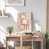 A beige framed poster with red text reading "YES YOU CAN" in a modern, minimalist font is displayed on a wooden desk in a bright, sunlit room. The setup includes a small plant, books, and a desk lamp, creating an inspiring and cozy workspace.