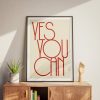 A beige framed poster with red text reading "YES YOU CAN" in a modern, minimalist font is displayed on a light wooden cabinet. The setup includes a small potted plant and a wooden hand sculpture, creating a warm and motivational space.